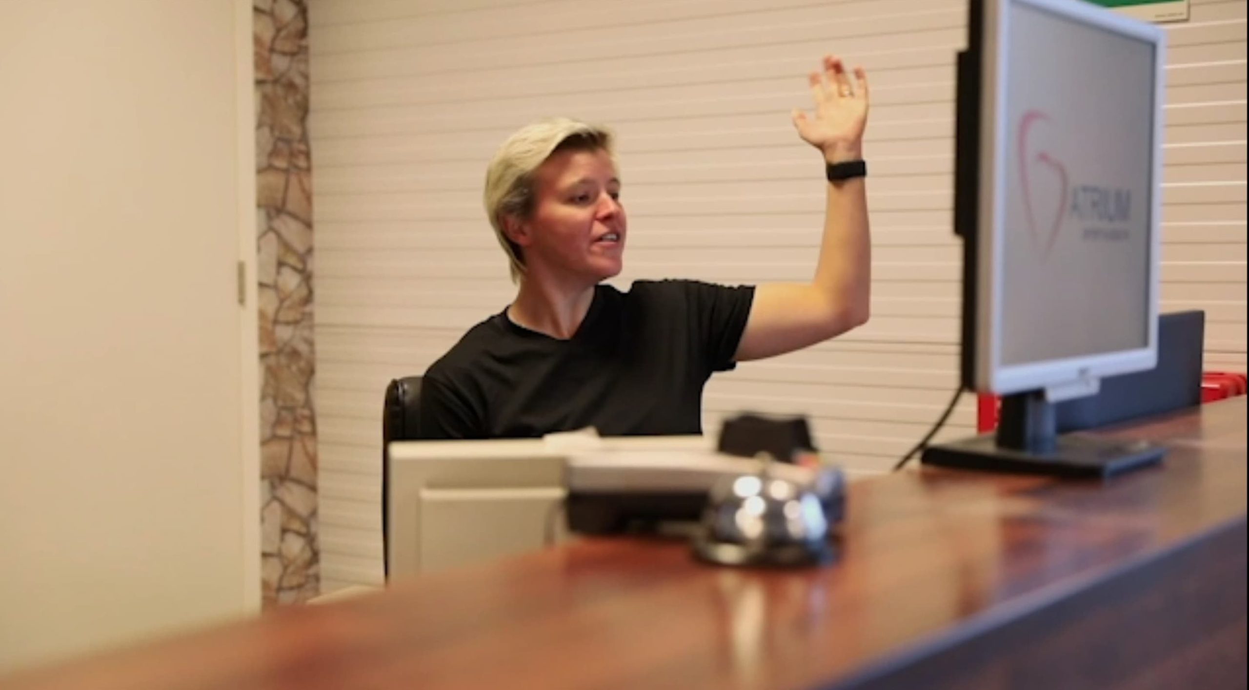 Een vriendelijke medewerkster van Atrium Ouddorp zwaait naar een klant vanaf de houten receptiebalie, met het logo van de sportschool zichtbaar op een computerscherm.