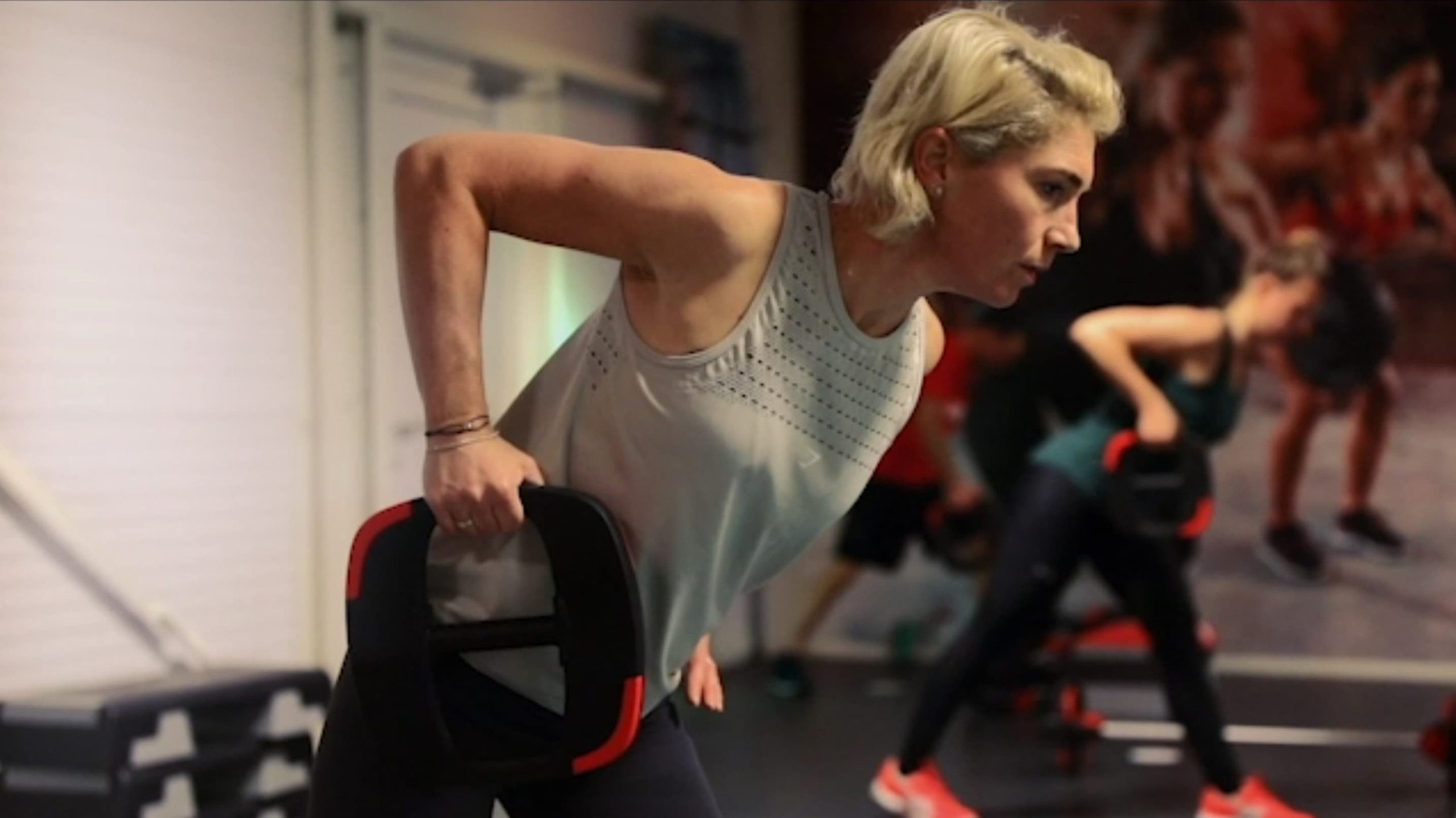 Een sportieve vrouw met kort blond haar voert een single-arm row oefening uit met een zwart-rode gewichtsschijf in een sportschool. Op de achtergrond zijn andere sporters en een fitnessmuurschildering te zien.