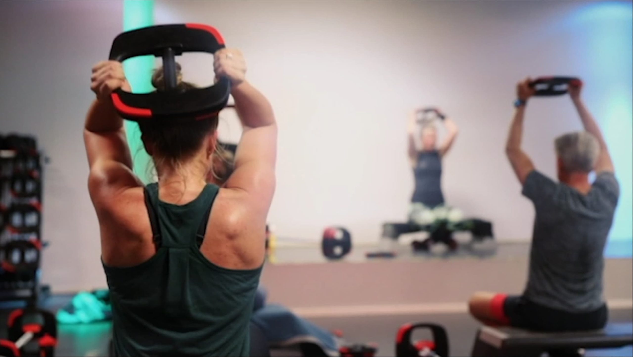 Een actieve groepsles bij Atrium Ouddorp waarbij deelnemers zittend op een stepbankje gewichtsschijven boven hun hoofd tillen voor een krachtoefening.